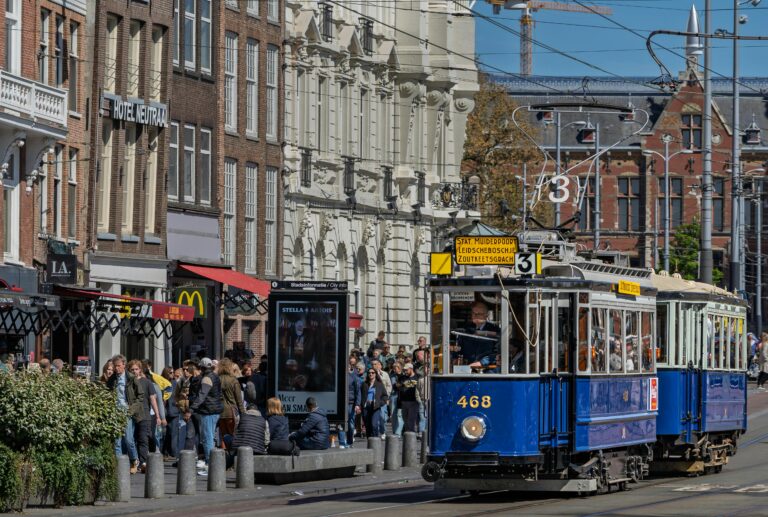 A historic blue tram rides through Amsterdam's busy street, capturing city life and classic architecture.